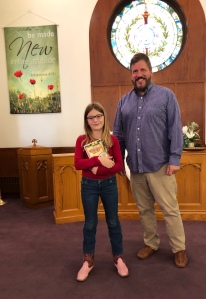 Two people, a young blonde girl in a red long-sleeved shirt and jeans with pink cowboy boots on the left and a tall white man with brown hair and short brown beard wearing a blue plaid shirt and khakis on the right, stand in a church sanctuary. The girl holds a bible.